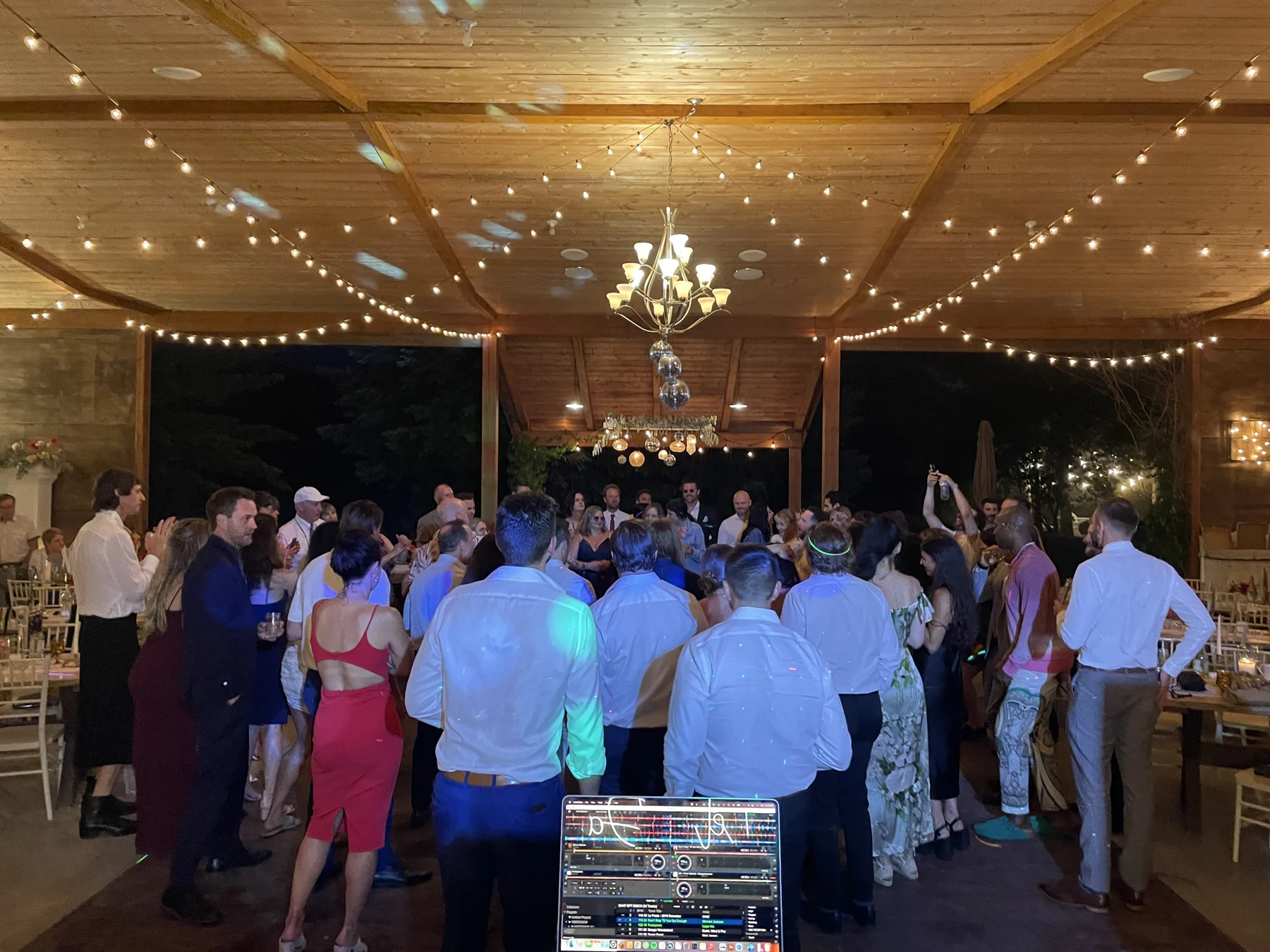 Wedding guests dancing together beneath chandeliers and string lights in a wooden pavilion